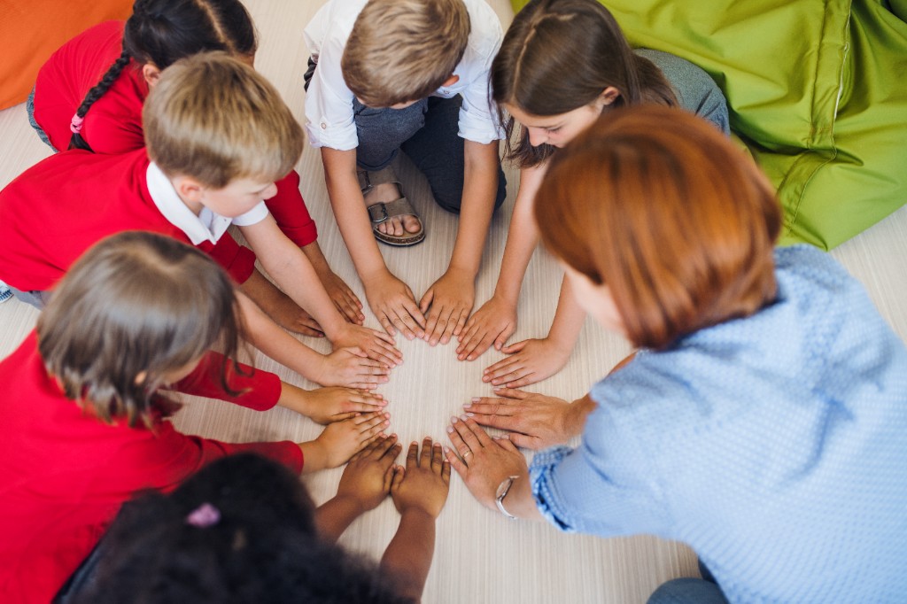 Overhead view of a diverse group of children and an adult sitting in a circle on the floor, placing their hands together in the centre to show unity and support.