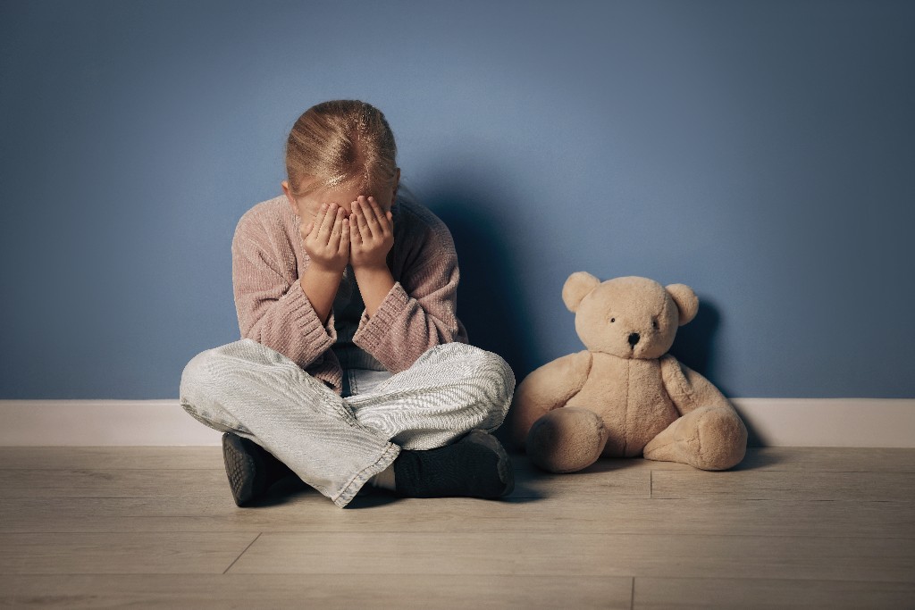 A young girl sitting on a wooden floor with her face buried in her hands, sitting next to a teddy bear against a blue wall, conveying a sense of sadness and loneliness.