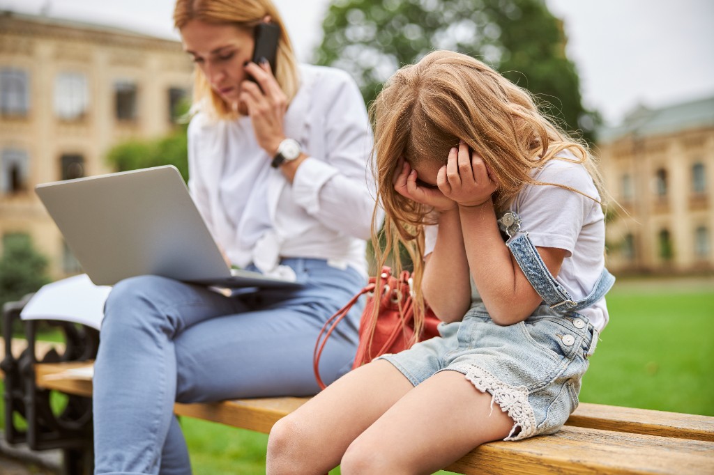 A young girl sits on a park bench with her head in her hands looking upset, while an adult sits next to her preoccupied with a laptop and phone call.