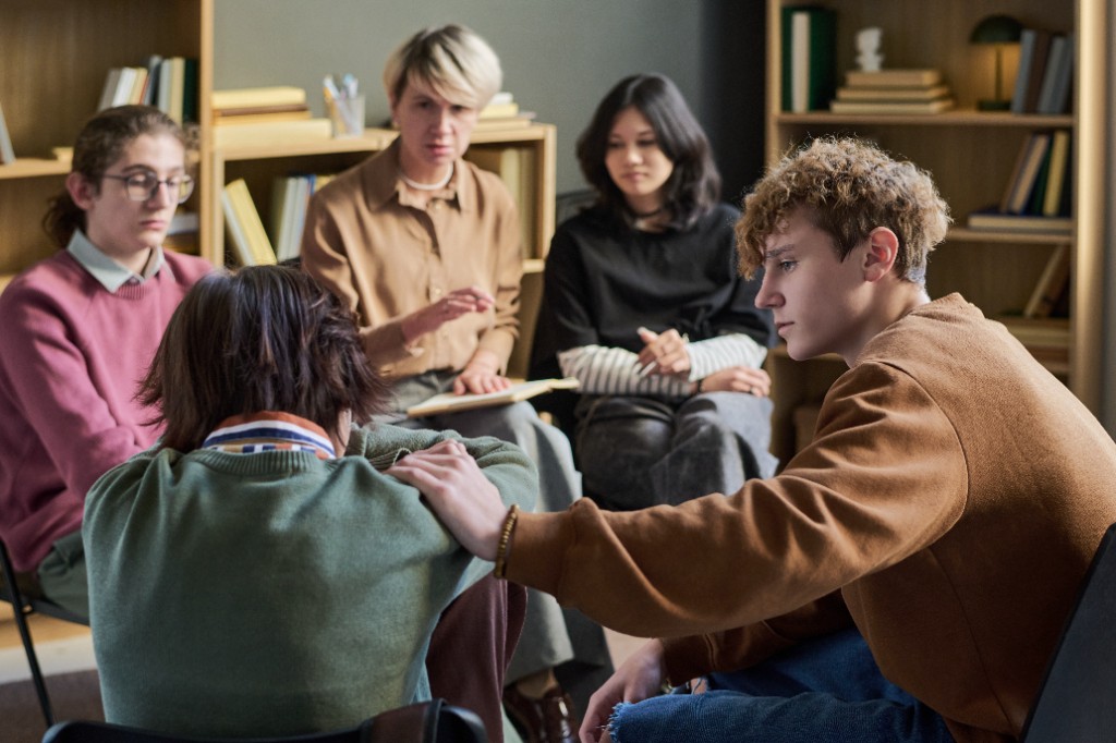 A group of teenagers and an adult facilitator sitting in a circle during a support session. One young man places a comforting hand on the shoulder of a peer who has their head bowed.